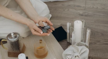 Personne tenant des pierres minérales colorées dans les mains, près d’une tasse, d’un bougeoir avec bougie et d’un récipient en verre sur une table en bois.