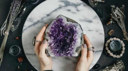 Porte-pierres violets en géode d’améthyste, tenue par deux mains sur socle marbre, entourée d’objets ésotériques.
