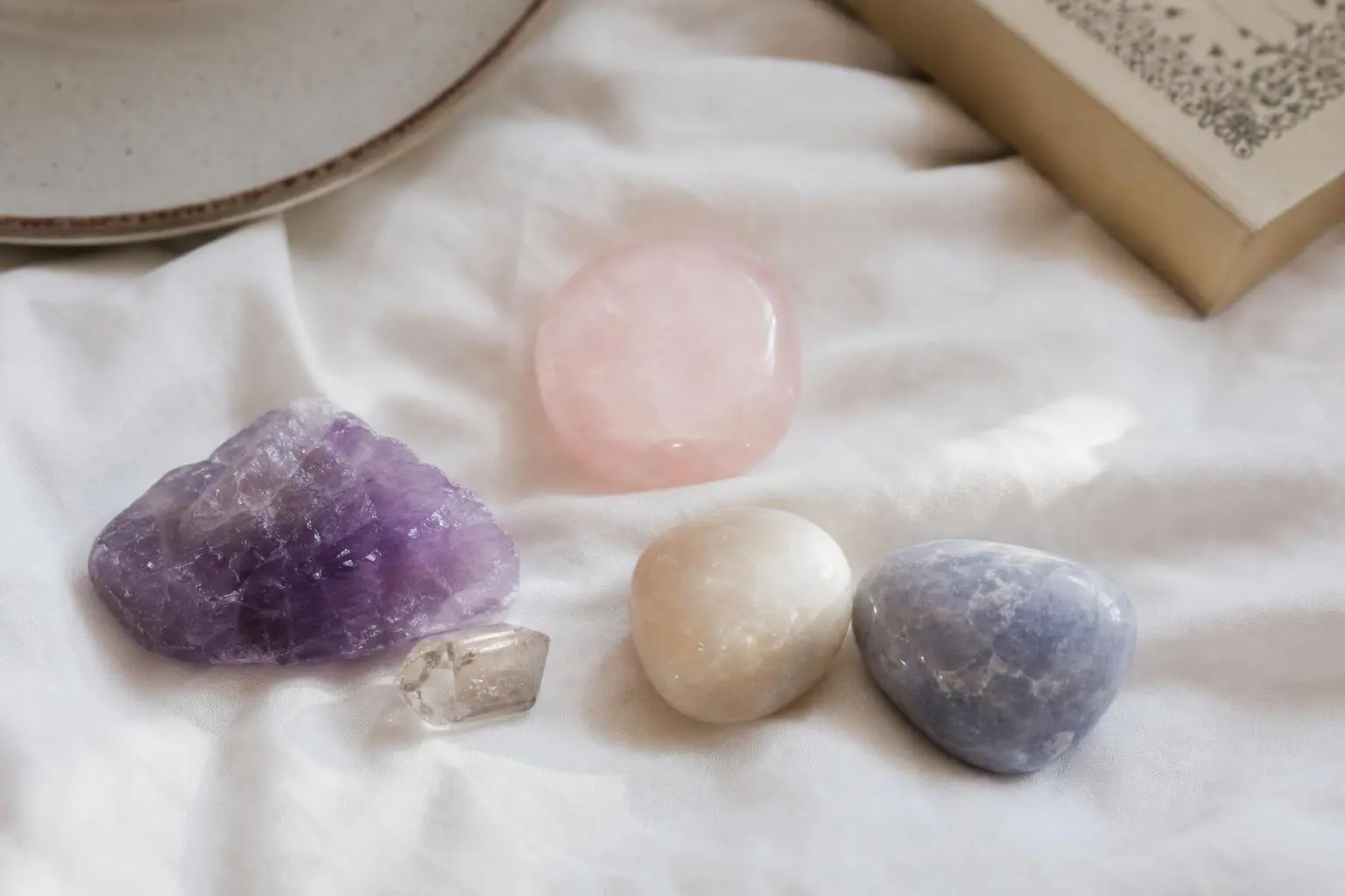 Plusieurs pierres roulées et cristaux colorés posés sur tissu, dont améthyste violette et quartz rose, avec livre en arrière-plan. peut-on dormir avec une pierre sous son oreiller ?